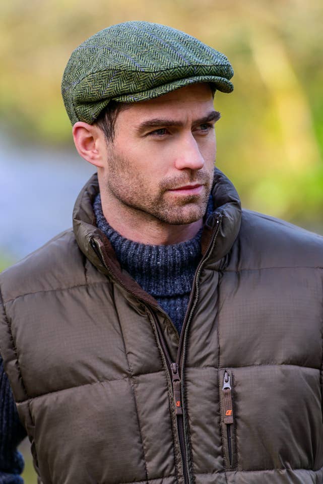 A man wearing an olive green herringbone flat cap, a brown puffer jacket, and a navy knit turtleneck, photographed outdoors with a softly blurred natural background.