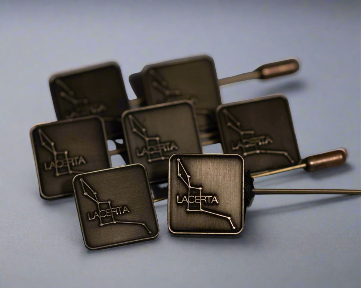 A collection of antique brass hat pins with the Lacerta logo die-struck on the top, displayed against a light blue background.