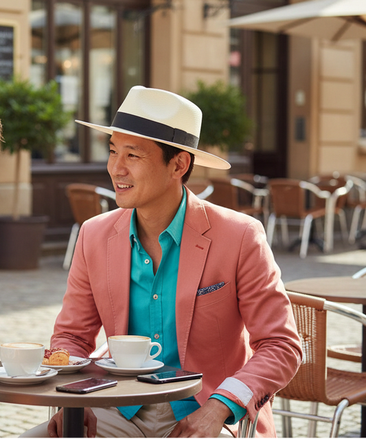 A man sitting at an outdoor cafe table wearing a light beige fedora hat with a black band, a pink blazer, and a turquoise shirt. Coffee cups and smartphones are on the table in front of him.
