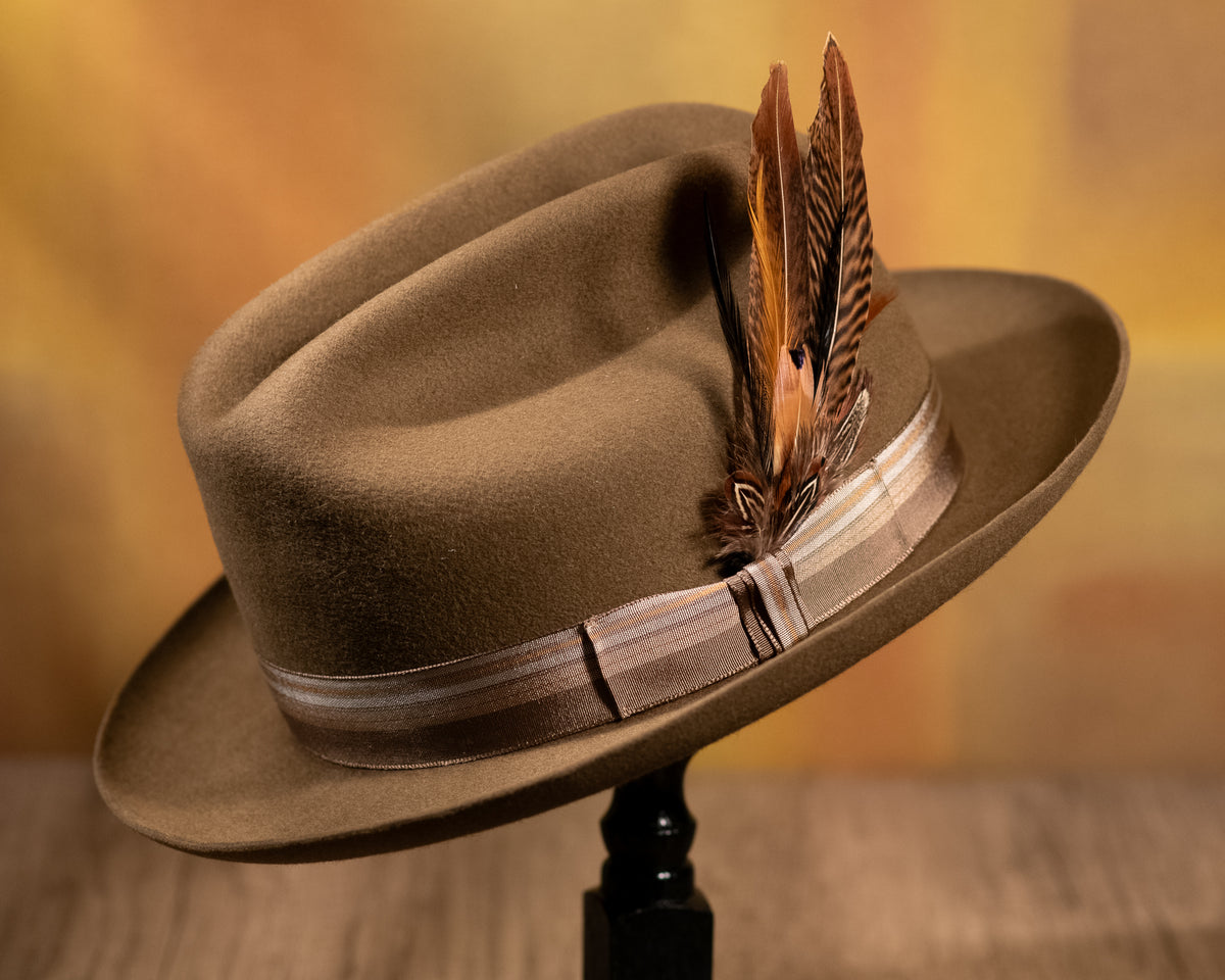 A dress weight beaver fur felt hat with a 2 1/2-inch upturn brim, featuring a striped taupe woven ribbon, a black leather sweatband, and a delicate feather accent.