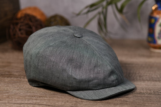 An olive textured newsboy cap with a curved brim, resting on a rustic wooden surface with a blurred background containing decorative yarn balls and a plant.