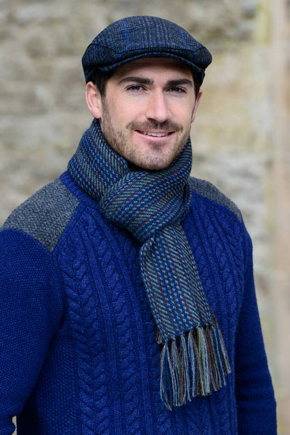 A man wearing a blue cable-knit sweater, a matching blue and gray scarf, and a flat cap, standing against a blurred stone wall.