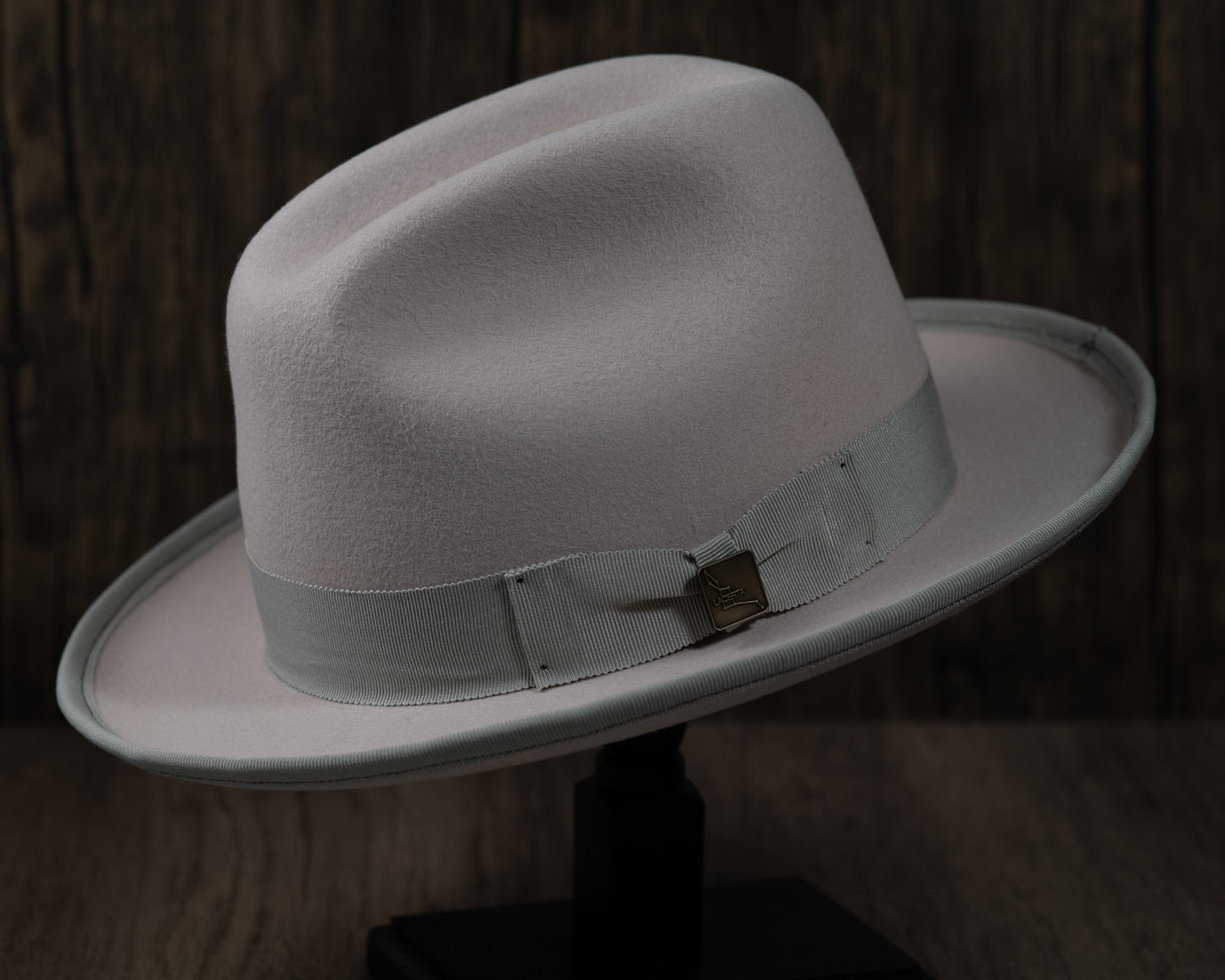 A Bone Rabbit Fur Fedora with a grosgrain ribbon and an upturned brim, displayed on a stand with a wooden background.