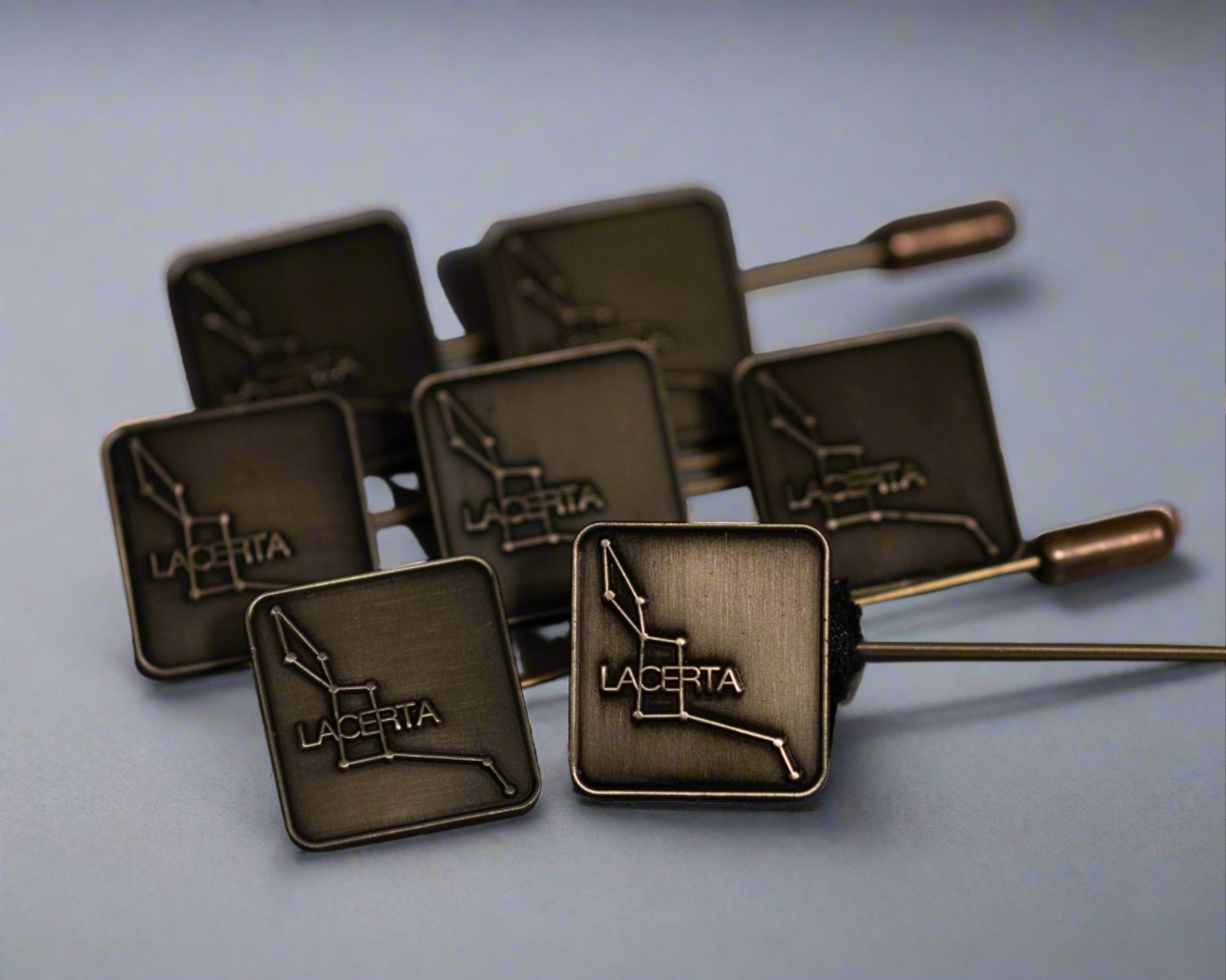 A collection of antique brass hat pins with the Lacerta logo die-struck on the top, displayed against a light blue background.