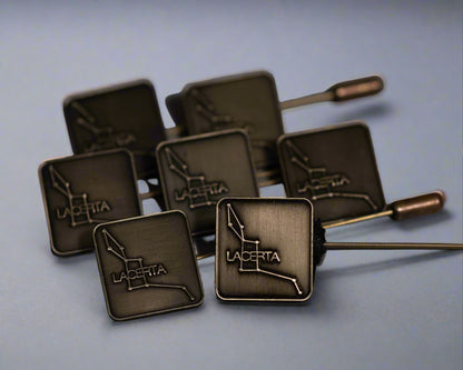 A collection of antique brass hat pins with the Lacerta logo die-struck on the top, displayed against a light blue background.