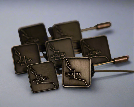 A collection of antique brass hat pins with the Lacerta logo die-struck on the top, displayed against a light blue background.