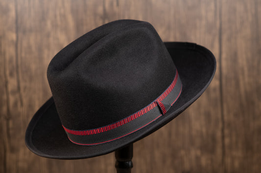 A black beaver fur fedora hat with a red band, displayed on a stand against a wooden background.