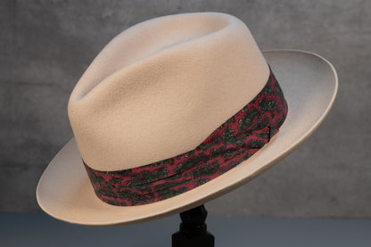 A bone white rabbit fur fedora with a red and green paisley hatband, displayed on a stand against a textured background.