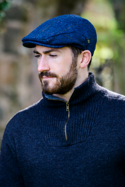 A man wearing a textured navy blue flat cap and a matching navy blue ribbed half-zip sweater, photographed outdoors with a blurred natural background.