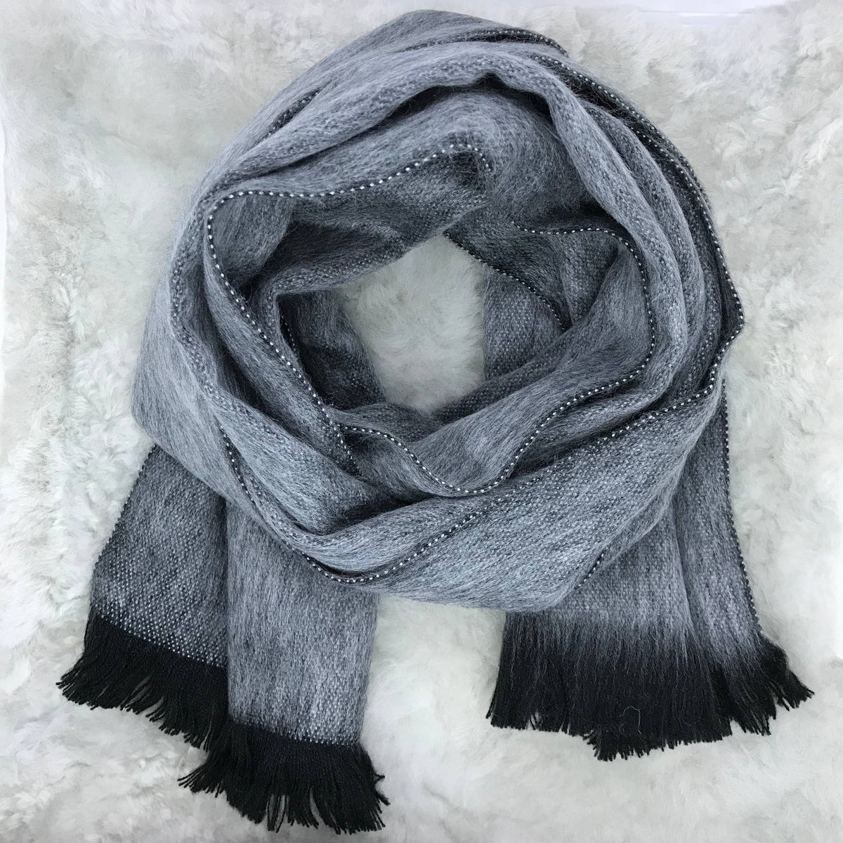 A gray and black gradient knit scarf with beaded edges and fringed ends, arranged in a loop on a white faux fur background.