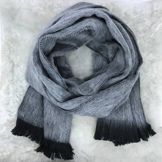 A gray and black gradient knit scarf with beaded edges and fringed ends, arranged in a loop on a white faux fur background.