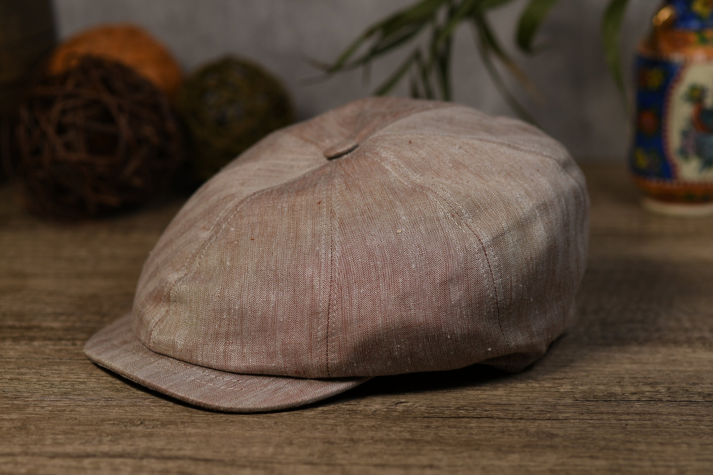 A textured, earthy brown linen flat cap with a curved brim, resting on a rustic wooden table with decorative background elements.