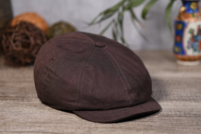 A dark brown eight-panel flat cap with a curved brim, resting on a rustic wooden surface with decorative props in the background.