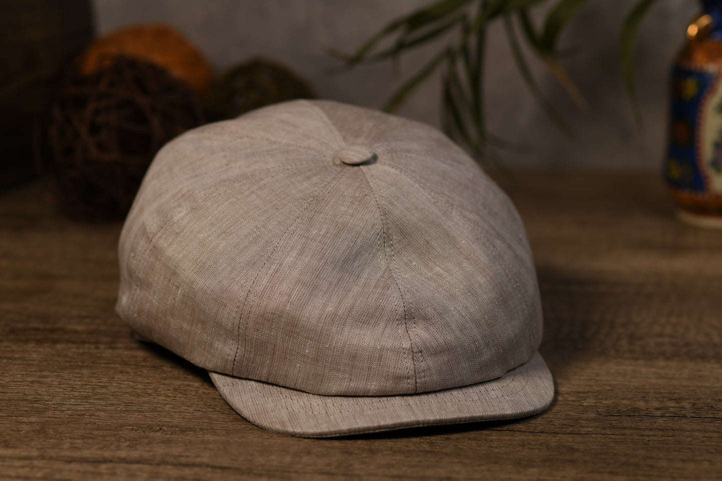 A light beige textured fabric newsboy cap with a curved brim, resting on a wooden surface with a blurred background.