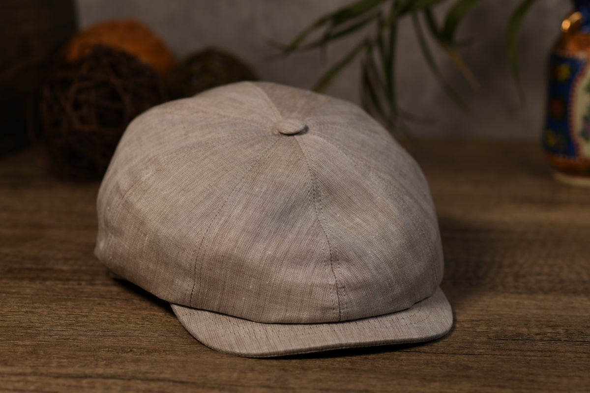 A light beige textured fabric newsboy cap with a curved brim, resting on a wooden surface with a blurred background.