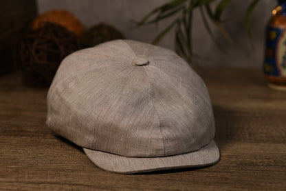 A light beige textured fabric newsboy cap with a curved brim, resting on a wooden surface with a blurred background.