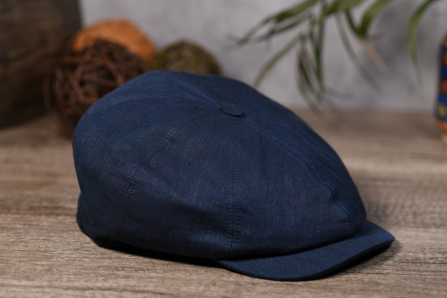 A dark navy blue flat cap made of textured fabric, resting on a rustic wooden surface with a softly blurred background containing decorative spheres and green leaves.