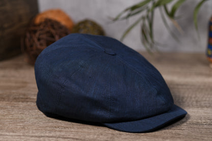 A dark navy blue flat cap made of textured fabric, resting on a rustic wooden surface with a softly blurred background containing decorative spheres and green leaves.