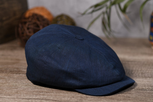 A dark navy blue flat cap made of textured fabric, resting on a rustic wooden surface with a softly blurred background containing decorative spheres and green leaves.