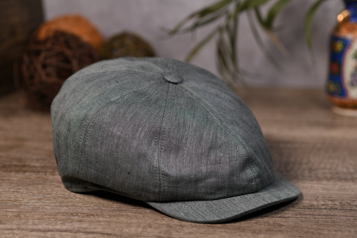 An olive textured newsboy cap with a curved brim, resting on a rustic wooden surface with a blurred background containing decorative yarn balls and a plant.