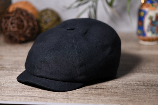 A black fabric newsboy cap with a curved brim, resting on a light wood tabletop with decorative woven balls and a colorful bottle in the blurred background.