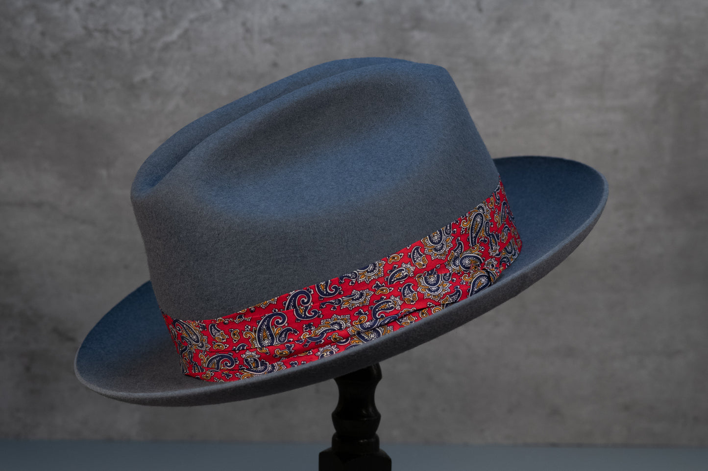 A steel grey rabbit fur felt hat with a red paisley hatband and a black leather sweatband, displayed on a stand against a grey background.